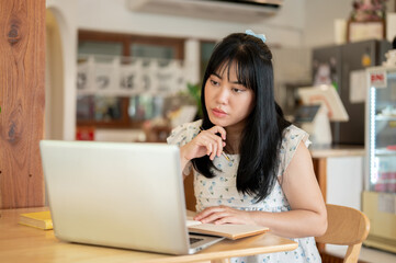A serious, thoughtful young Asian woman is focusing on her work, working remotely at a coffee shop.