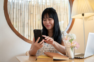 A happy, charming young Asian woman relaxing in a coffee shop and using her smartphone.