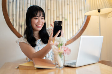 A cheerful young Asian woman is talking on a video call on her smartphone with her friend.