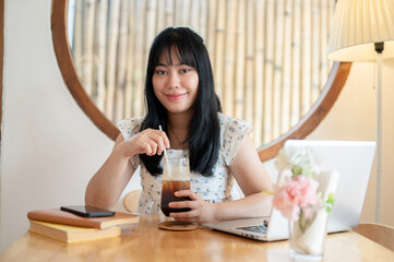 A beautiful Asian woman, dressed enjoys her iced coffee while sitting at a table in a coffee shop.
