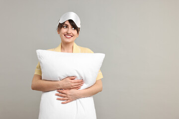 Happy woman in pyjama and sleep mask holding pillow on grey background, space for text