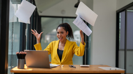 A businesswoman in a yellow jacket expressing frustration and throwing papers into the air at her office workspace.