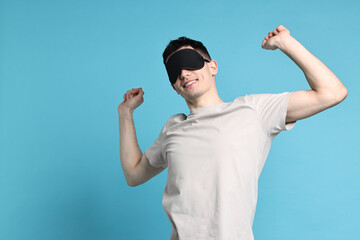 Happy man in pyjama and sleep mask stretching on light blue background, space for text