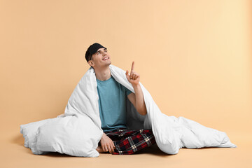 Happy man in pyjama and sleep mask wrapped in blanket on beige background