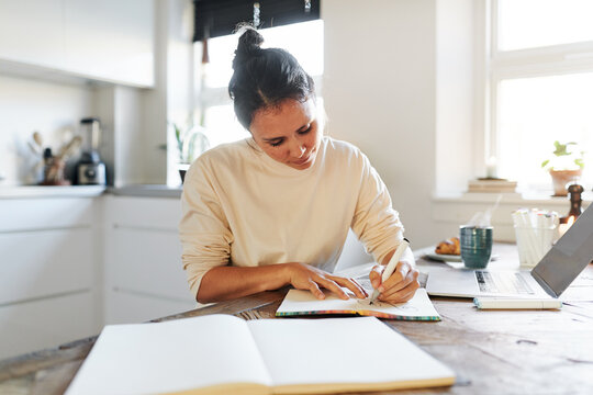 A woman sits focused in her kitchen and sketching a design in a book. On the table is an open laptop and there are several open notebooks