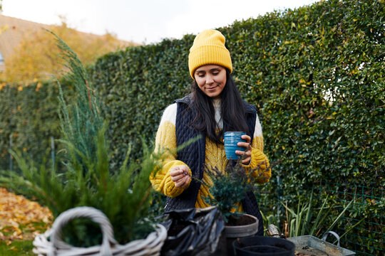 A smiling woman stands in her garden and enjoys a cup of coffee while doing garden work. She wears a yellow hat that matches her shirt