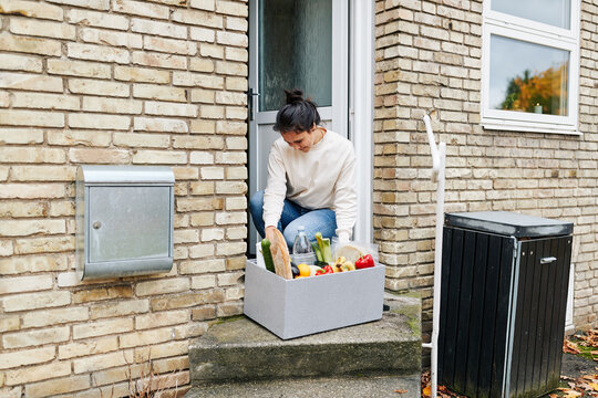 Woman looking through a grocery delivery box. Which has just been delivered to her front door outside. In the box there are fresh organic vegetables, and freshly baked bread.