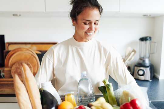 A smiling woman stands in her kitchen. In her arms she has a box full of fresh groceries. She happily looks down at her goods. In the box there are fresh organic vegetables, and freshly baked bread.