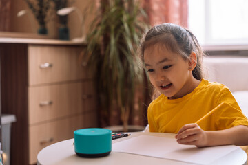 Child draws at the table and talks to a blue smart speaker.