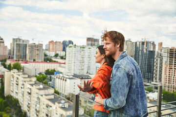 Fototapeta na wymiar A man and woman stand on a balcony, gazing at a sprawling city below them under the night sky