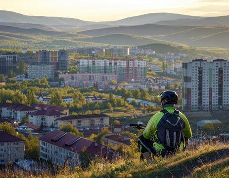 The silhouette of a young man in a helmet with a backpack on a mountain bike against the background of an evening sunset. View of the evening city at the foot of the mountain. Mountain biking. An acti