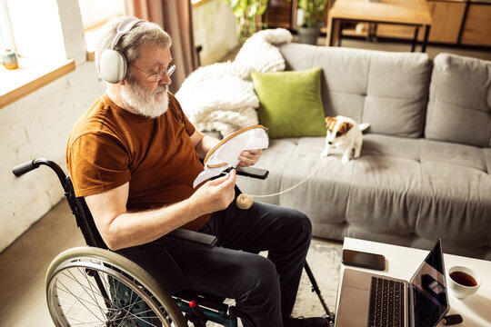 Elderly man sitting on wheelchair in living room at home on daytime and embroidery. Leisure and relaxing activity. Concept of healthcare, lifestyle, wellness, comfort, empowerment