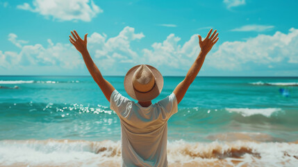 Back view of a traveler wearing a hat with arms up in the air standing at the beach in front of the sea in background during summertime holidays