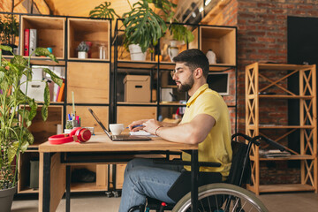 Young man, employee sitting on wheelchair and working remotely on laptop at home, Remote job,...