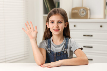 Happy little girl waving hello at white table indoors © New Africa