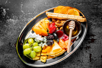 An elegant cheese and fruit platter with various cheeses, fresh fruits, honey, and breadsticks, beautifully presented on a dark background