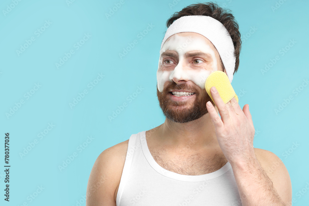 Wall mural Man with headband washing his face using sponge on light blue background, space for text