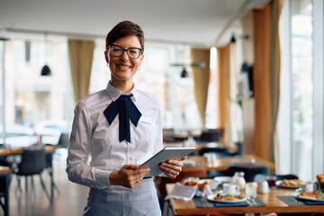 Happy waitress with touchpad at hotel restaurant looking at camera. - obrazy, fototapety, plakaty