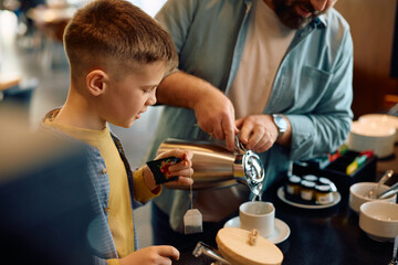 Smiling boy and his father making tea while having breakfast in hotel restaurant.