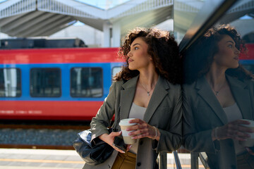 Gazing into the distance, she stands by the station’s glass barrier, balancing coffee and phone,...