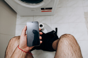 A man is sitting on the toilet with a phone in his hand. Guy's morning routine