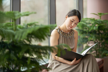Reading, plants and woman in home with book to relax with glamour, comfort and elegant fashion. Eco, green decor and Asian girl in house with files, reference and cool carbon neutral interior design