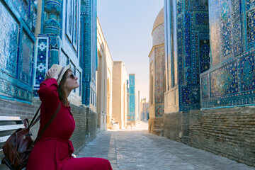 Woman with hat and red dress relaxing on the bench and enjoying the beauty of the ancient Shah-i-Zinda complex, Samarkand, Uzbekistan