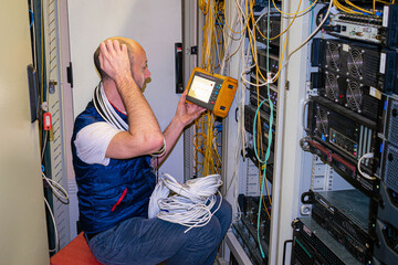 A man wrapped in wires works in a server room. A technician measures the level of an optical signal while sitting in a data center.
