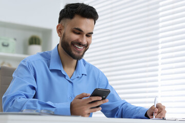 Happy young man using smartphone while working at white table in office, low angle view
