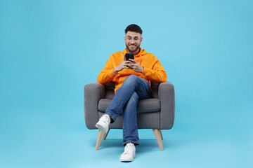 Happy young man using smartphone on armchair against light blue background