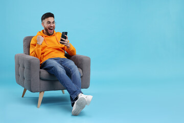 Happy young man using smartphone on armchair against light blue background, space for text