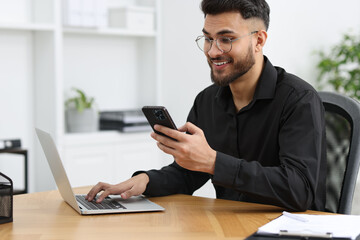 Handsome young man using smartphone while working with laptop at wooden table in office