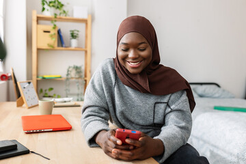 Happy gen z young black woman in muslim headscarf using cell phone at home. Social media, youth...