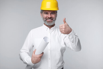 Architect in hard hat holding draft and showing thumb up on grey background