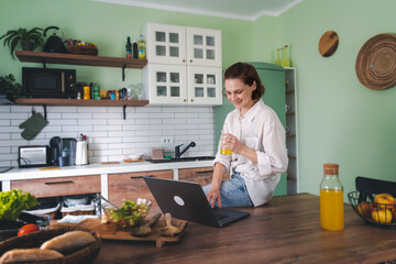 Young cheerful woman using laptop at home in kitchen, working from home