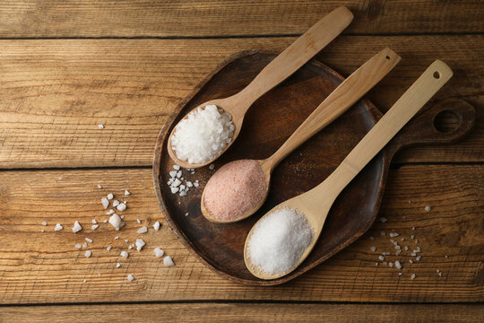 Different natural salt in spoons on wooden table, flat lay. Space for text
