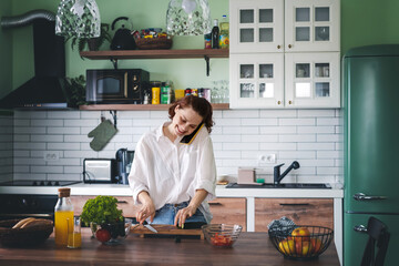 Young cheerful young woman talking on mobile phone while cooking at the kitchen.
