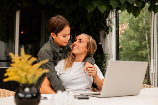 Smiling woman sitting with laptop while looking at son hugging from behind in back yard