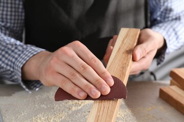 Man polishing wooden plank with sandpaper at grey table, closeup - obrazy, fototapety, plakaty