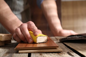 Man polishing wooden plank with sandpaper at table indoors, closeup - obrazy, fototapety, plakaty