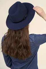 Close-up shot of a young woman in a dark blue felt fedora hat with black strap. The girl with long brown hair is wearing blue jeans shirt and a blue hat and posing on a beige background. Back view.