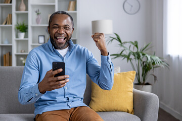 Portrait of an enthusiastic mature man celebrating a victory with his mobile phone, sitting...