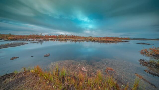 Fast-moving clouds move in the sky above the shallow river with forest-covered banks flowing through the Finnish tundra. A timelapse video.