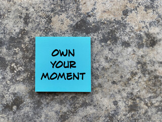 Motivational and inspirational wording. OWN YOUR MOMENT written on a notepad. With blurred styled background.