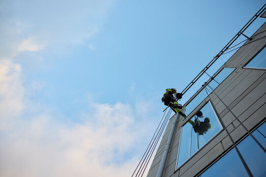Low angle view of male rope access worker hanging on glass building under blue sky