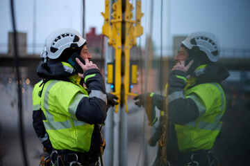 Male rope access worker talking on phone while standing with coworker outside window of building