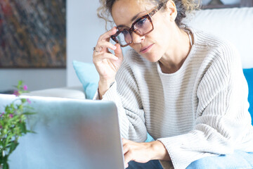 Thoughtful woman holding laptop on laps, pondering ideas or tasks, sitting on couch at home, dreamy young female touching glasses and looking the display, lost in thoughts, waiting for message online