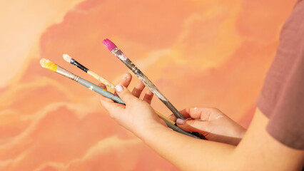 A girl holding three painting brushes on the background of an artwork on the wall in pink and peach fuzz colors