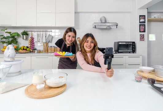 Two women enjoy in a fun cooking or baking activity in a modern kitchen. Capturing video or photo with a smartphone on a selfie stick while holding a handful of colorful baking tools or decoration.