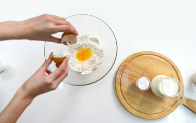 Closeup hand cracking an egg into a bowl of flour, preparing to bake. Baking powder and a jug of fresh milk are on kitchen counter. Top view